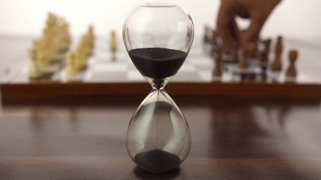 Closeup shot of hands playing chess next to a glass sand clock during leisure time. Checkered board with black and white chess pieces  a strategy board game played by two smart players - white back...