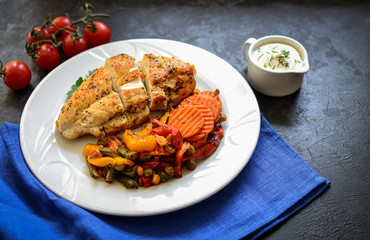 Baked chicken breast with vegetables. Chicken breast, asparagus, carrots, in a white plate. Dark background. Free space for text. Blue linen napkin.