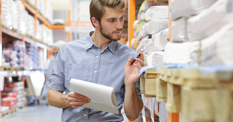 Young man shopping or working in a hardware warehouse standing checking supplies on his tablet.