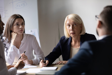 Focused diverse businesspeople sit at desk brainstorm discuss business project at meeting together, concentrated colleagues consider ideas cooperate at office briefing, collaboration, teamwork concept