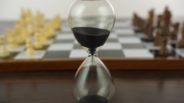 Closeup shot of black and white chess pieces on a checkered board - white background. Racking focus shot of a chessboard with wooden chess pieces  next to a glass sand clock - a strategy board game 