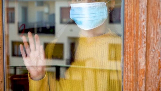 Hopeful young caucasian south american girl with dark hair yellow shirt ank blue mask on looking outside her window and posing her hand on it during covid-19 lockdown