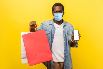 Online shopping and delivery on quarantine. Portrait of young man with medical mask holding packages and showing smartphone and looking with smile. indoor studio shot isolated on yellow background