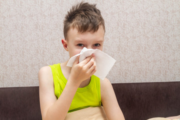 A teenage European boy is lying on the bed . The child is ill. The boy blows his nose on a white napkin. Rhinitis.