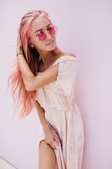 Cute caucasian girl playing with her pink hair and smiling. Stylish young woman in summer dress and sunglasses posing on white background.