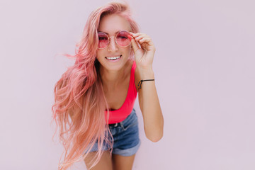 Interested girl with light-pink hair touching her sunglasses. Studio shot of attractive curly woman in trendy summer outfit smiling on white background.