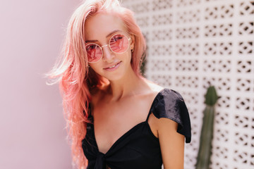 Portrait of winsome caucasian lady in pink sunglasses posing near cactus. Fashionable white female model in black dress standing in front of exotic plant.