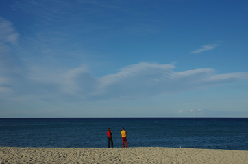 Obraz premium A man with red trousers and yellow shirt next to a woman with black trousers and red shirt look at the sea