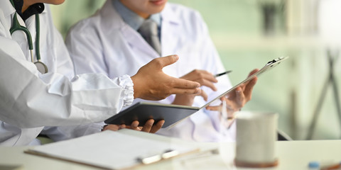 Cropped image of two smart men working as doctor holding a computer tablet and clipboard while discussing and sitting together at the doctor working desk over orderly workplace as background.