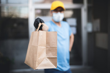 Delivery guy with protective mask and gloves holding box / bag with groceries in front of a building.