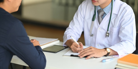 Cropped image of Young smart doctor diagnosing and giving advice to his patient while sitting together at the doctor working desk over comfortable clinic as background.