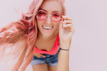Close-up portrait of adorable stylish girl playfully touching her pink glasses. Sensual female model in denim shorts having fun during indoor photoshoot.