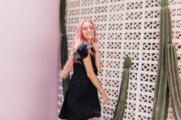 Charming caucasian fair-haired woman posing in black dress near cactus. Outdoor shot of pretty female model with pink hair wears sunglasses.