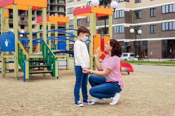 Fototapeta na wymiar family mother and son at the playground. the child wears a protective medical mask during an epidemic coronary virus or flu. personal protective equipment. Mother gives hand antiseptic to her baby