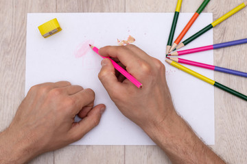 a man's hands draw with a pink pencil on a white sheet next to which are a yellow sharpener,...