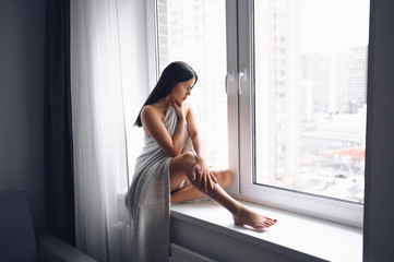 Beautiful bored slender brunette woman sitting next to window windowsill under warm grey blanket at home. Self isolation quarantine during Corona virus pandemic. COVID19 stay home save lives concept