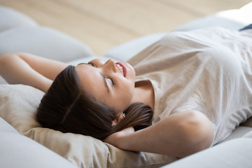 Close up satisfied happy woman with hands behind head relaxing and lying on comfortable sofa during break at home. Smiling attractive girl resting after house work done closed eyes, daydreaming.