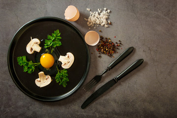 Fresh, raw chicken eggs with mushrooms and herbs, a fork and a knife on a black dish on a gray stone background, top view. Ingredients for cooking, ideas for the kitchen, minimalistic style.
