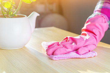 The hands of a young woman in a pink rubber glove holds a soft rag, wipes the table from dust. House cleaning, washing surfaces in the living room on a sunny day, the concept of cleanliness.
