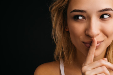 Naklejka premium Young cheerful playful blonde woman posing on camera. Close up and cut view of girl holding finger on lips and look left. Hush symbol. Isolated over black background.