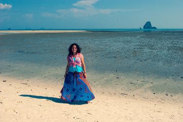 Young beautiful girl in a dress stands on the white sand by the sea at low tide. European appearance and black hair. The weather is sunny. Blue sky with clouds. Tinted. Horizontal.