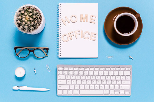 Flat lay of modern workspace with keyboard, coffee cup, notepad, pen, eyeglasses and cactus on blue background. Letters saying Home Office. Top view