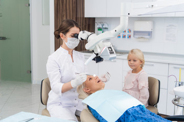 A blond woman having her teeth checked at the dentist's office, with her daughter waiting for her