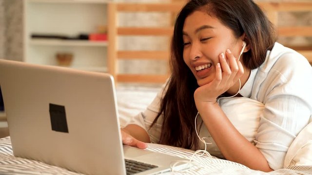 Happy young Asian girl in casual clothing lying down on bed while making a video call with laptop computer in bedroom at home. Video conferencing technology concept.