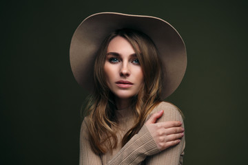 A young attractive blonde woman in a beige sweater, fedora hat and brown leather trousers poses on a dark green olive background. Studio portrait of a beautiful girl.