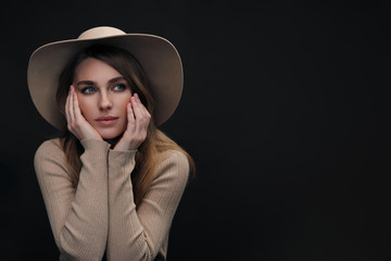 A young sensual sexy woman with a beautiful face and clear natural skin poses in the studio against a black background. Beige slinky sweater and hat with fields.