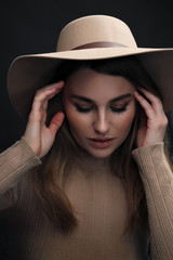 A young sensual sexy woman with a beautiful face and clear natural skin poses in the studio against a black background. Beige slinky sweater and hat with fields.