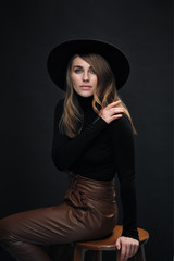 Portrait of young beautiful blond woman with light makeup and blue eyes touching her face. Dark background. Black tight sweater and hat with fields.