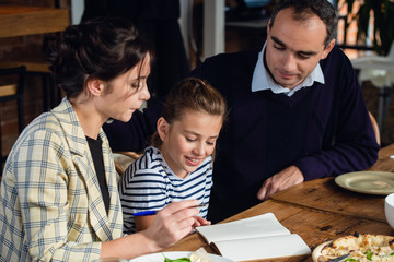 Naklejka premium A girl doing her homework at the kitchen table with her parents being around