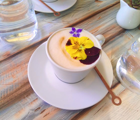 Сup of cappuccino decorated with flowers on a wooden table and glasses with water. Coffee and flowers in the rays of sun.