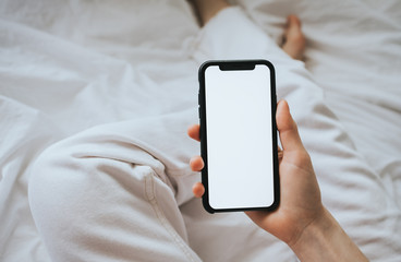 Woman holding a smartphone with a white screen, sitting on the bed at home on a sunny day. Mock-up