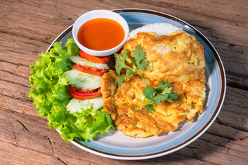 Omelette with minced pork topped with rice, served with chilli sauce, cucumber, tomato, arranged in a beautiful dish, placed on a wooden table.