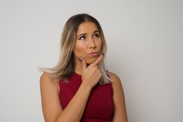 Isolated portrait of stylish young European woman with hand under chin and looking sideways with doubtful and skeptical expression, worry and doubt. Standing indoors over gray background.