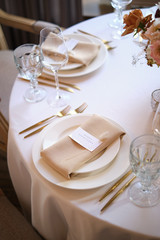 Luxury cozy autumn wedding table decoration in the restaurant. Fresh and dried flowers, roses, carnations. Beautiful table setting: golden appliances, fork and knife, calligraphy guest seating card.