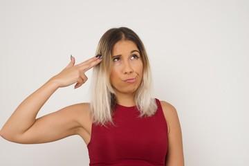 Fototapeta premium Unhappy blonde young female imitates gun shoot makes suicide gesture keeps two fingers on temples has puzzled expression dressed in casual clothes stands indoors.