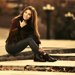 Happy young fashion woman in leather jacket sitting on sidewalk