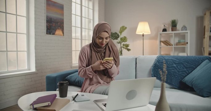 Young muslim girl wearing traditional hijab scarf looking at her smartphone. Successful female freelance manager or university student working at home or office 4k footage