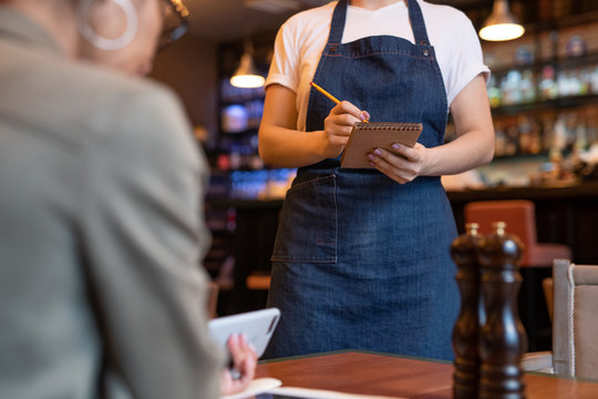 Young waitress in apron and white t-shirt writing don order of client in notepad