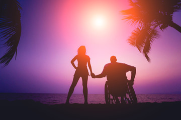 Disabled man in a wheelchair with his wife on the beach. Silhouettes at sunset