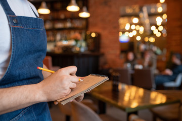 Hand of young waiter in denim apron holding pencil over page of notepad