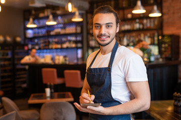 Young smiling waiter in workwear making notes in notepad while working