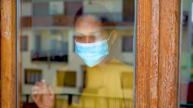 Hopeful young caucasian south american girl with dark hair yellow shirt ank blue mask on looking outside her window during covid-19 lockdown