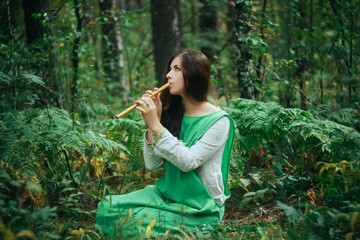 A medieval woman in a green dress plays on a wooden flute sitting in fern bushes. A girl in a gloomy forest plays music on a handmade pipe. Young witch in a twilight fantasy forest. © Михаил Гута
