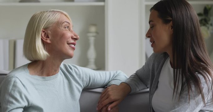 Head shot close up side view smiling middle aged woman relaxing on couch with happy grown up daughter, talking about life moments. Bonding affectionate two generations family chatting in living room.