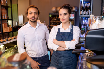 Two young colleagues in white shirts and denim aprons standing by workplace