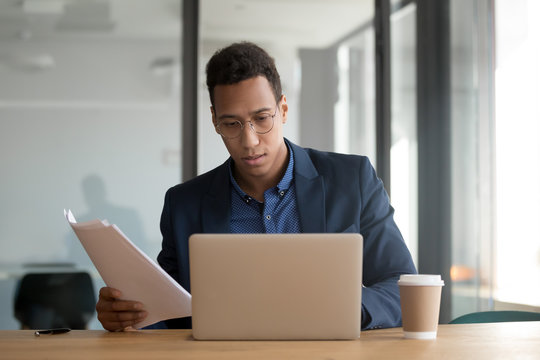 Focused busy african ethnicity businessman wearing suit and glasses checking documents read analyzing statistics sales data work results do paperwork using computer working in modern office concept